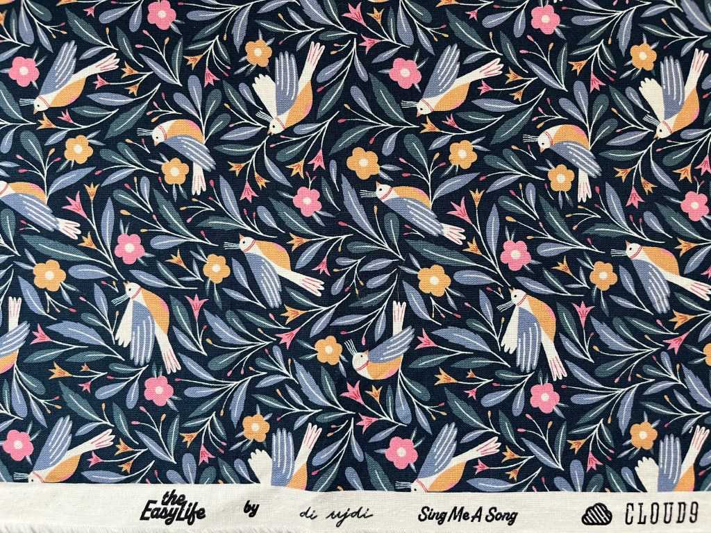 The image features a close up of fabric printed with blue birds with a tan belly on a dark blue background. There are pink and tan flowers and blue leaves. The text on the selvedge of the fabric reads The Easy Life by di ujdi Sing Me A Song Cloud9.