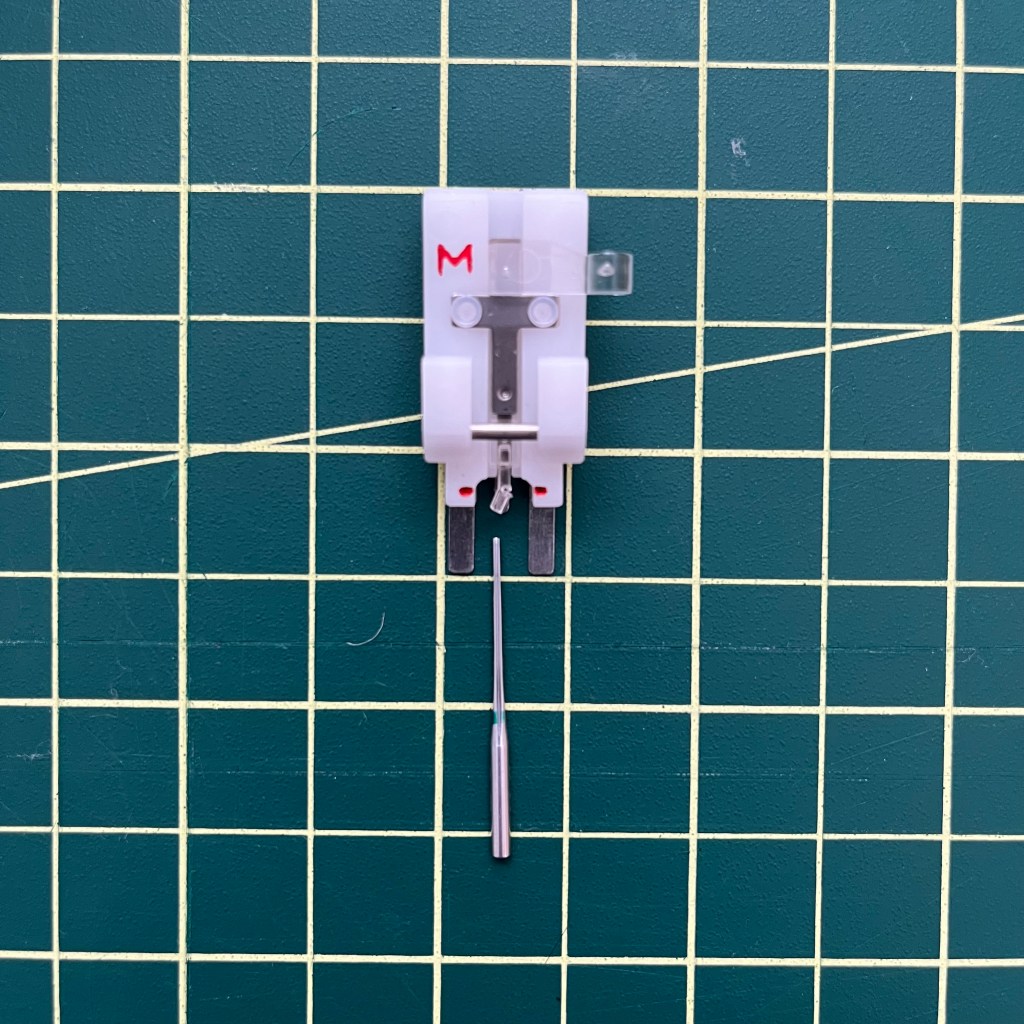 A photo of a white sewing presser foot for sewing a button onto a garment on a gridded green cutting mat. A small plastic piece on the foot is broken, as is the tip of a sewing machine needle sitting next to the foot.